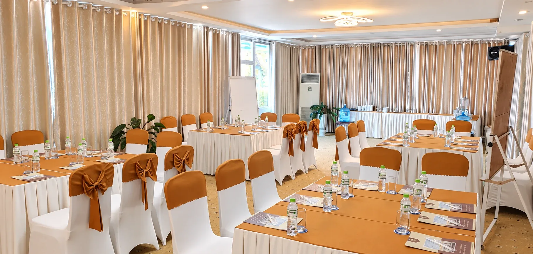 Bàn nhóm phủ khăn với nước chai và tài liệu, flipchart và bàn trà nước phía cuối phòng